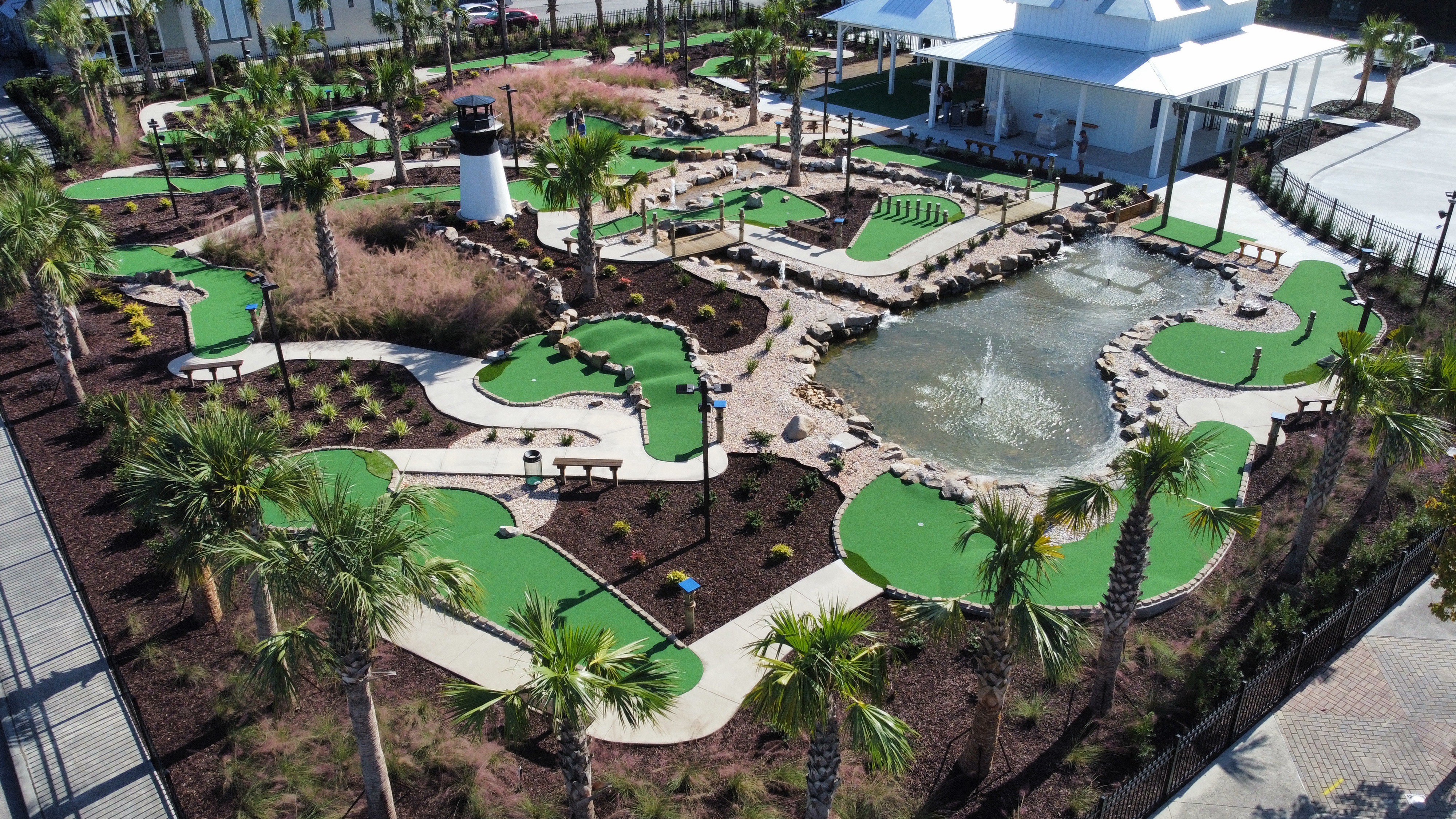 Aerial view of the entire mini golf course showing 18 holes with bright green surfaces, themed obstacles, water features, and palm trees throughout the grounds.