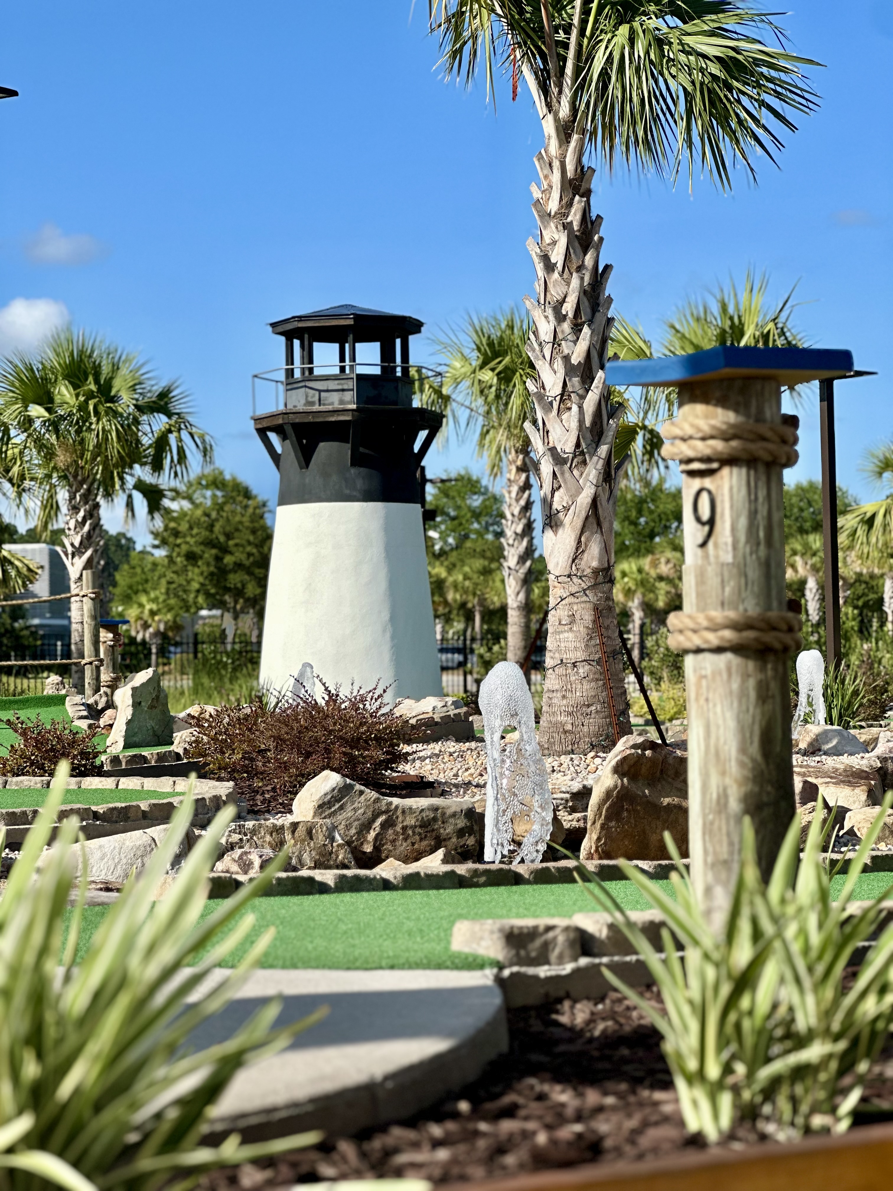 A single mini golf hole featuring a lighthouse-themed obstacle with a white and black tower, hole marker showing 9, and surrounding tropical vegetation.