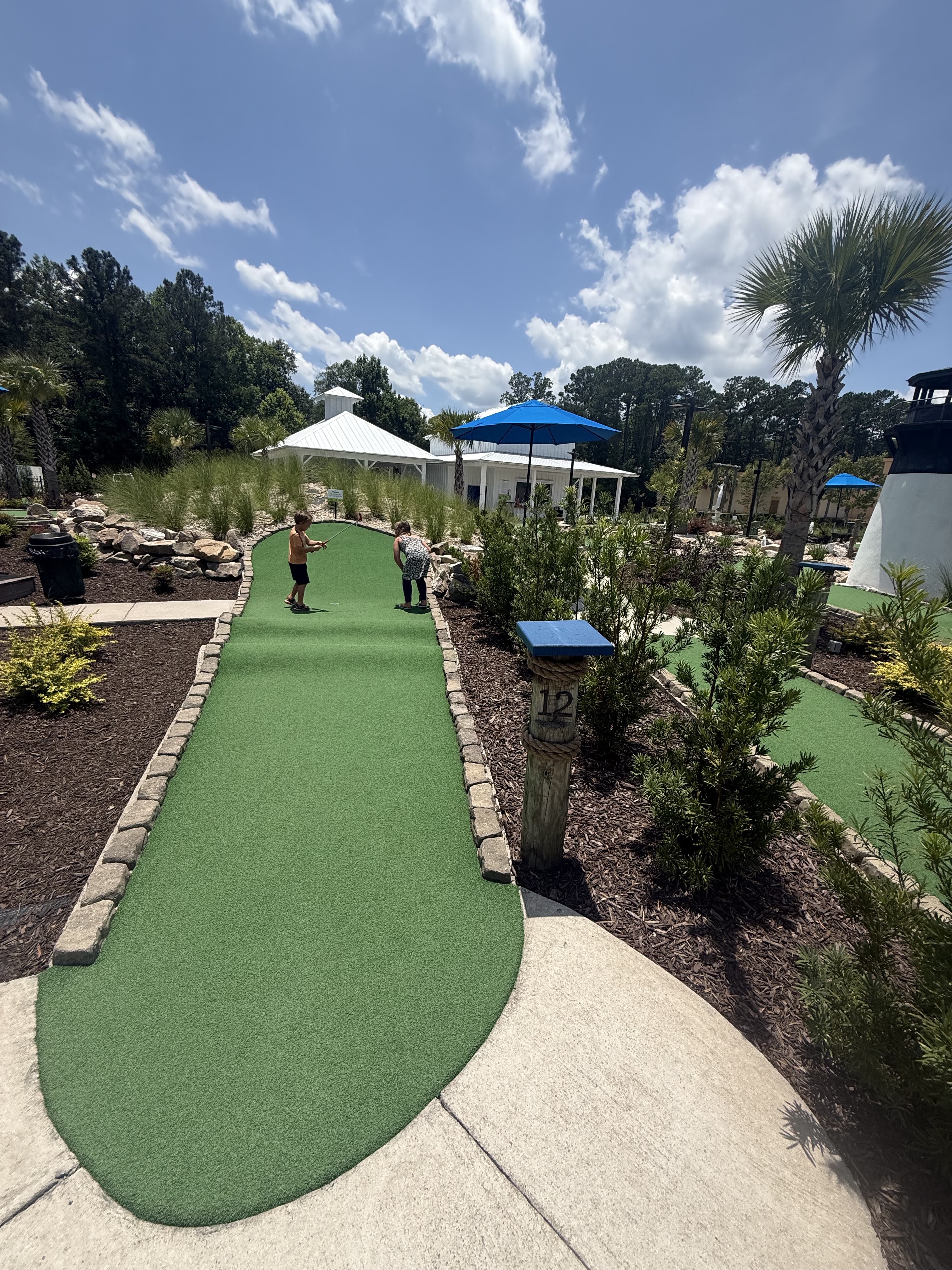 Players teeing off on a mini golf hole with shaded pavilions visible in the background and well-maintained green surface with hole marker 12.