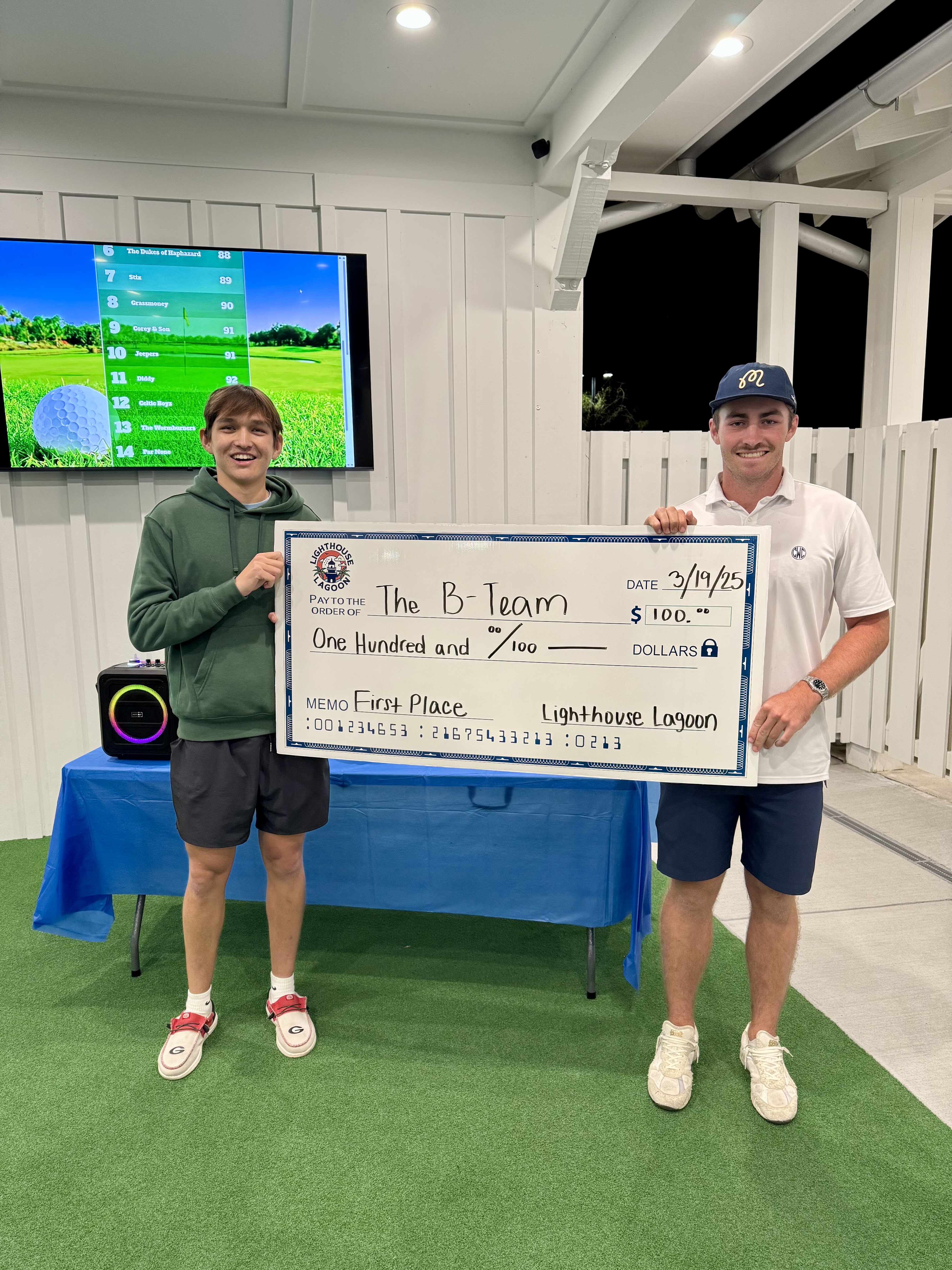 Two people holding a ceremonial first-place check for $100 in an interior space with a scorecard displayed on a TV screen above and green putting surface visible.