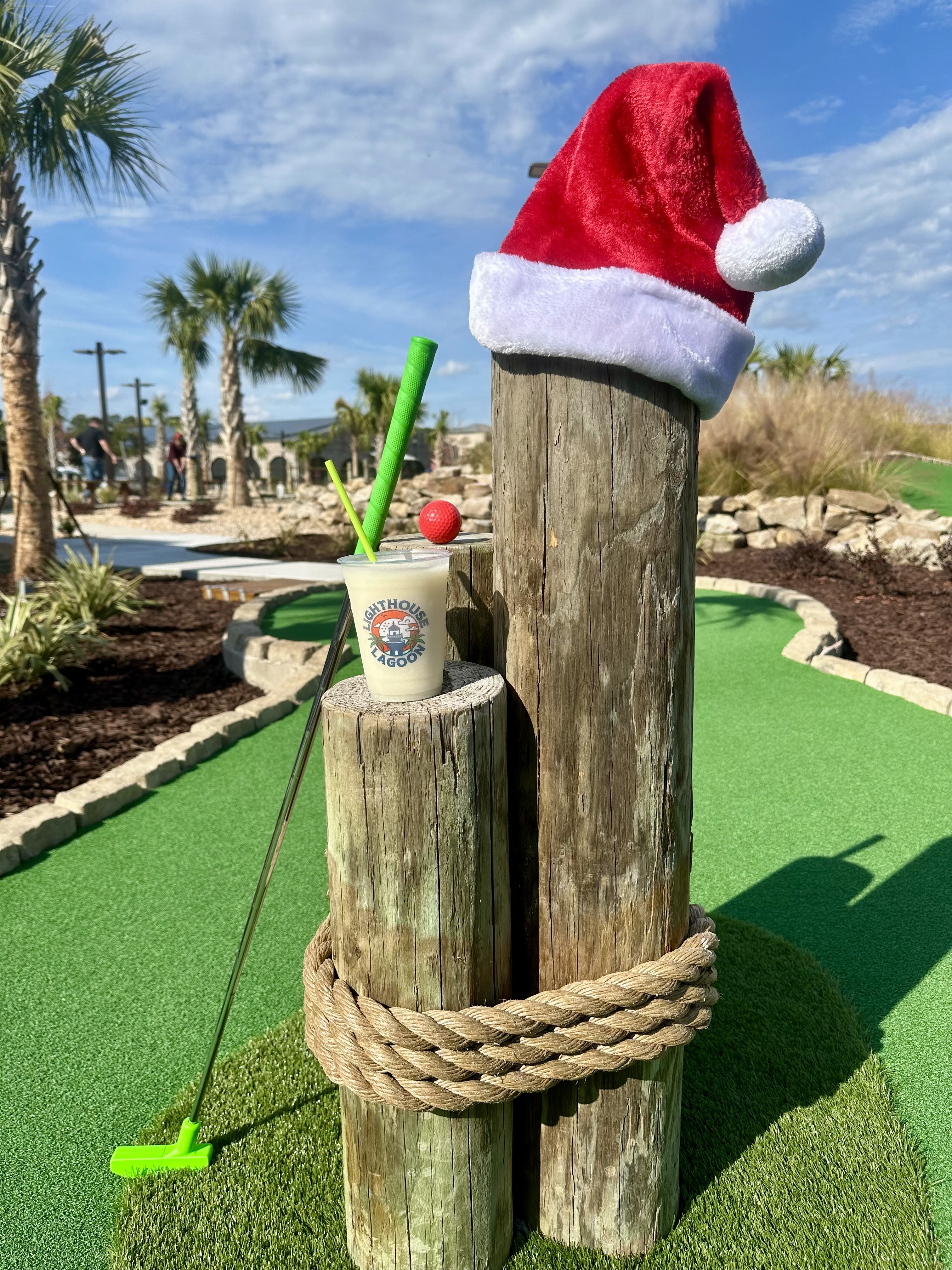 A themed mini golf hole with a wooden post decorated with a Santa hat, holding a drink cup with the Lighthouse Lagoon logo, festive decorations, and hole marker visible on the green surface.