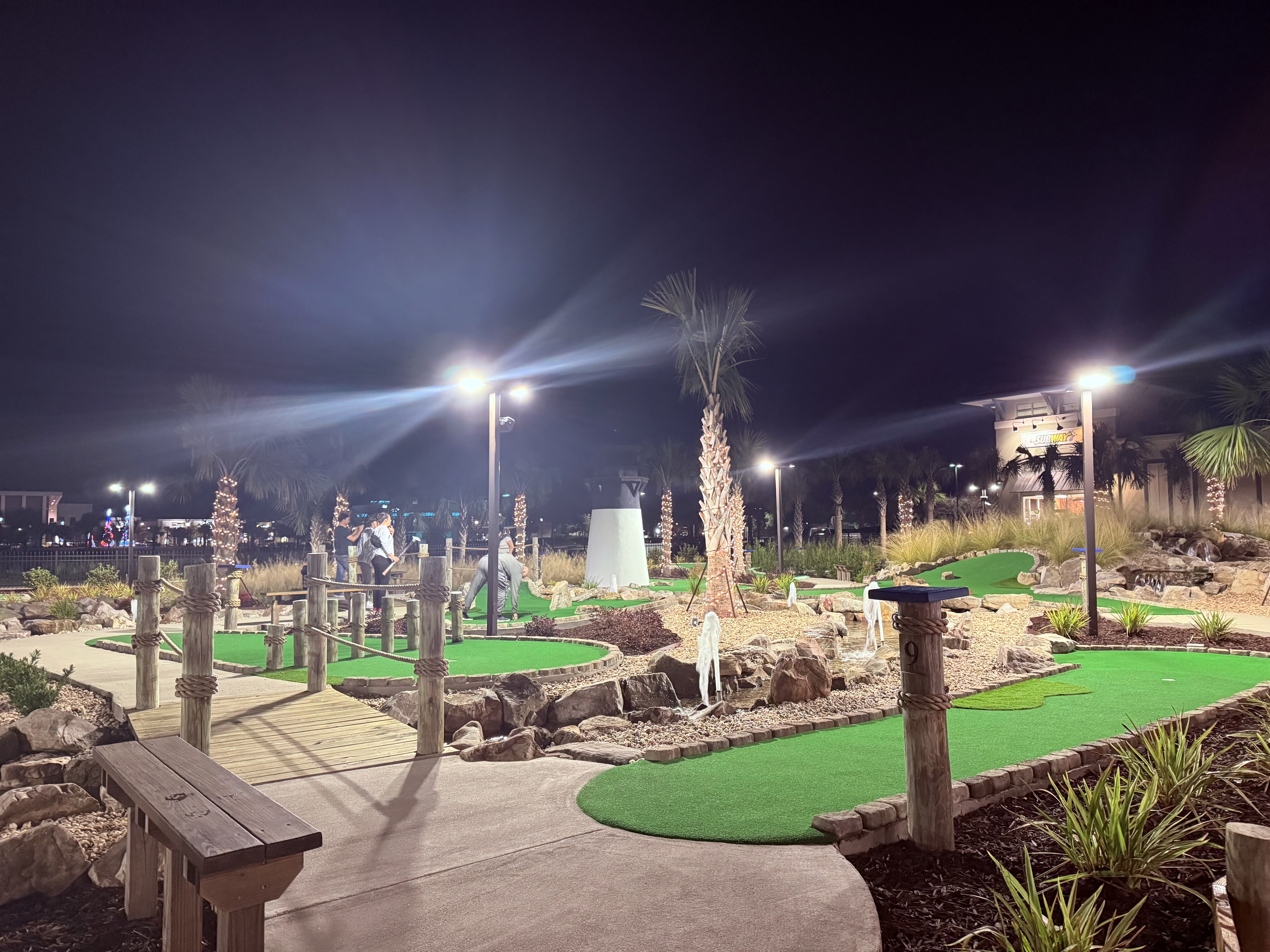 Night-time view of the mini golf course illuminated with bright LED lighting, showing multiple holes with green surfaces, wooden railings, and palm trees throughout.