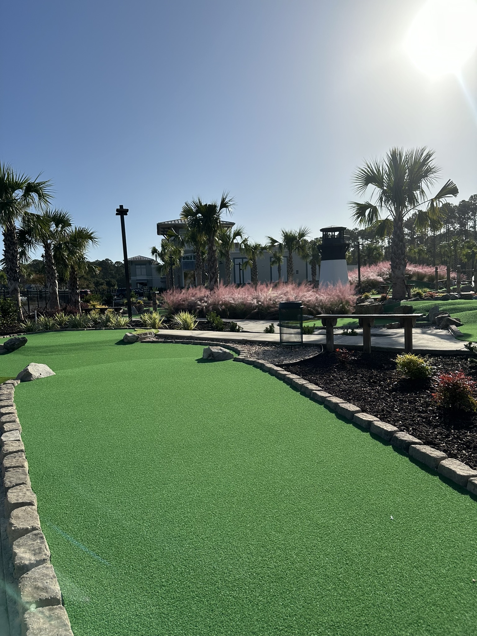 Wide-angle view of a mini golf hole showing the green putting surface, wooden bridge obstacle over water, palm trees, themed architectural structures in the background, and clear daylight.